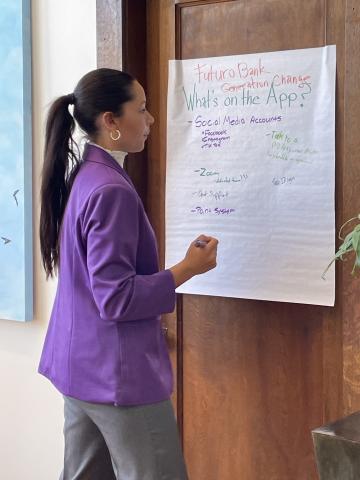 Female college student wearing a purple blazer and writing on a large sheet of white paper posted to the wall.